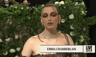Met Gala 2024 gif. Closeup of Emma Chamberlains full dark eyeshadow completely covering her eyelids and up to her eyebrows and dark brown lipstick, adding to the gothic-style look of her dress. Two thin curtain-style strands swoop down from the crown of her head to tuck behind her ears.