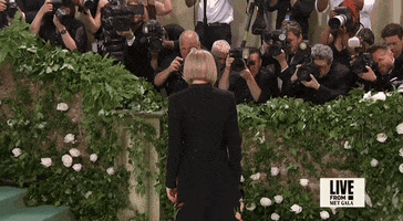 Met Gala 2024 gif. Anna Wintour turns and poses for photos, placing her hands in her pockets. She's wearing a white floor-length gown with a black floor-length coat buttoned in the middle over it. The coat has red and yellow tulips with green stems as appliqués adding pops of color and texture to the coat.
