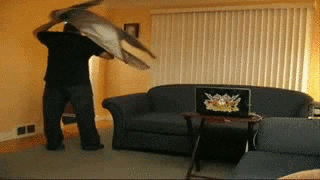 Video gif. Man stands in a living room putting his coat on. A woman jumps out behind vertical window blinds and scares him. The man freaks out, running into the wall, falling backwards in the armrest of the sofa and onto the ground.