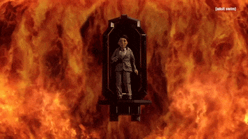 TV gif. A puppet from the Shivering Truth wears a suit stands on a floating platform as it's engulfed in the flames that blaze around him.