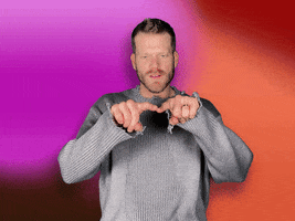 Video gif. Scott Hoying of Pentatonix draws a giant heart with their fingers and then points at us with a sweet expression against a pink-orange gradient background.