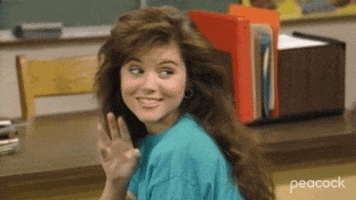 TV gif. Tiffani Thiessen as Kelly Kapowski on Saved By The Bell looks over her shoulder as she sits at her desk in school. She has a flirtatious smile and she wiggles her fingers to playful say hello. 