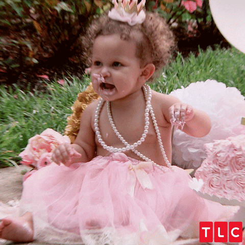 Video gif. Toddler in a pink tutu and tiara with a double string of pearls hanging down her bare chest seated on a lawn filled with pink paper party decorations. She has pink cake all over her face as she continues to put more cake in her mouth on loop, opening and closing her mouth like she's babbling at someone offscreen.