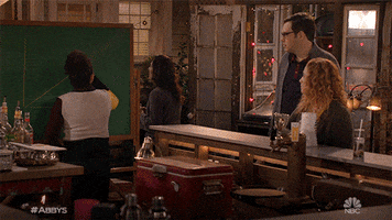 TV gif. Three bar patrons from Abby's watch a woman draw a line graph on a chalkboard. The woman draws an upward diagonal line, continuing the line's upward path off the board before twirling around.