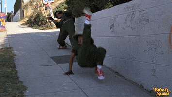Video gif. Woman in a green jumpsuit and safari hat breakdances down a sidewalk on a hill on all fours, her back to the ground. Behind her, a man in a matching jumpsuit bends his legs like noodles and moves his torso in a circular motion.