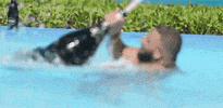 Celebrity gif. DJ Khaled in a swimming pool wrestling with a bottle of champagne half his size, which explodes in an arc over his head.
