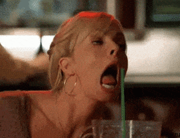 Video gif. A blonde woman acting drunk with her mouth open and tongue out, trying to reach for a green straw.