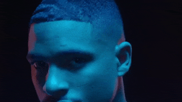 Music video gif. Arin Ray in A Seat music video is washed over in blue and orange light. He moves around, locking eyes with us as he moves his head around.