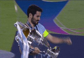 Sports gif. Rodri of Manchester City holds up the giant UEFA Champions League trophy and looks at us as he kisses and struts proudly across the field. 