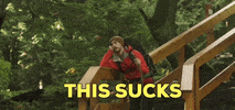 Video gif. Man leans on the railing of wooden stairs in the middle of a forest, wearing camping gear and holding a large stick as a cane. We zoom in on his face as he turns his head to look at us with a bored expression and says, “This sucks.”