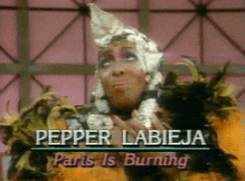 Movie gif. Pepper Labieja from Paris is Burning fancily adorns a neon orange and black boa and a glittering silver headpiece She clasps her hands together and looks hopefully towards the sky.