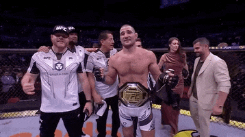 Sports gif. We zoom down to Sean Strickland's UFC championship belt which he is wearing behind his waist. Others stand in the ring with him, looking like they are getting ready to take a picture. 