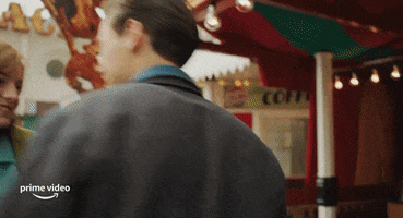 Movie gif. Harry Styles as Tom in "My Policeman," stands outside of a carnival game cheering and celebrating with his arms open wide, then kisses the girl next to him on the cheek.