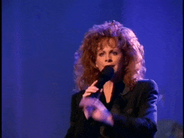 Celebrity gif. Singing on stage, Reba McEntire raises a hand as if to say, “stop.”