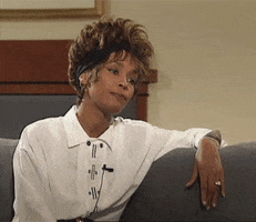 Celebrity gif. Whitney Houston leans her arm over the back of a couch, looking deeply bored, blinking.
