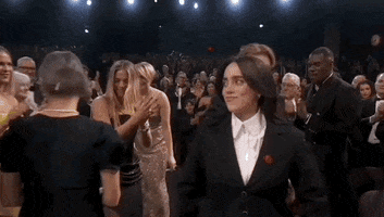 Oscars 2024 GIF. Billie Eilish and Finneas O'Connell walk towards the stage to accept their award for Best Song before Eilish spontaneously diverges to run and hug America Ferrera. 