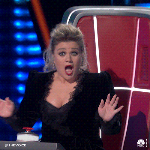 Reality TV gif. Kelly Clarkson as a judge in The Voice quickly stands up from her chair, hands raised in excitement with her jaw dropped and eyes wide as if astounded.