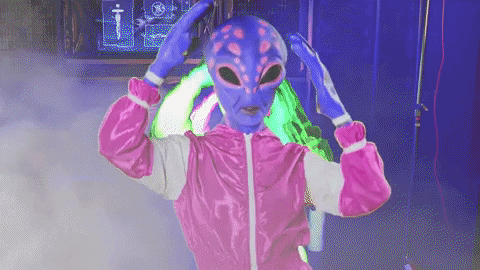 Video gif. Person dressed as a purple alien in a pink jumpsuit voguing in a smoky colorful atmosphere.