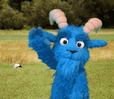 Video gif. Blauer Bock Blue Goat waves hello from a field of grass.