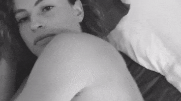 Video gif. A woman is lying in bed topless and we see her head, neck, and collarbones. She looks sultry as she tosses us a look over her shoulder.