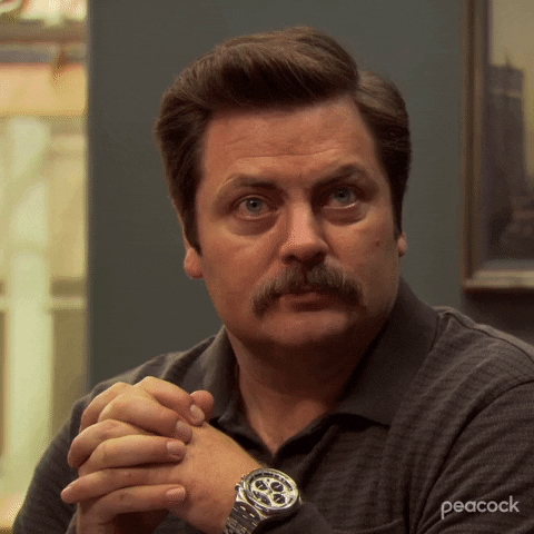 Parks and Recreation gif. Nick Offerman as Ron squints and scoffs and partially flips open his clasped hands as if to say "whatever."