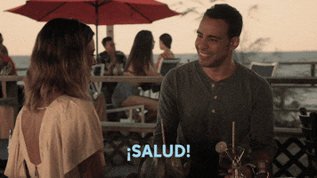 TV gif. Victor Rasuk as Daniel and Nathalie Kelley as Noa in The Baker and the Beauty tip their drinks to each other as he smiles at her and they say, "Salud! Salud! Salud" back and forth to each other.