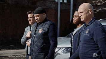 TV gif. A group of firemen from Chicago Fire are standing together with their heads bowed. One does the sign of the cross as the others look distraught.