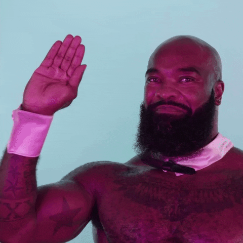 Video gif. A bald man with a large black beard wears only a shirt collar and bowtie plus shirt cuffs. He smiles broadly and waves stiffly to us.