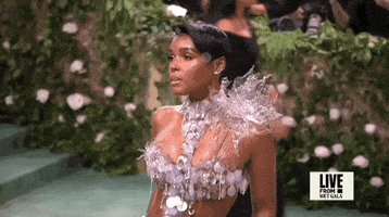 Met Gala 2024 gif. Slow motion clip panning down Janelle Monae's dress featuring a mostly see-through dress accented with holographic disks across her chest and down through the skirt. She wears a bursting silver poinsettia-like design at the top of one shoulder.