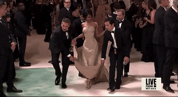 Met Gala 2024 gif. Tyla takes short steps in high heels as four assistants carry her train. She's wearing a sand-colored sculpted Balmain mermaid-style dress that's completely fitted through her angled waistline and hips. The fabric through the skirt is stiff and structured, giving the appearance of waxed fabric. 