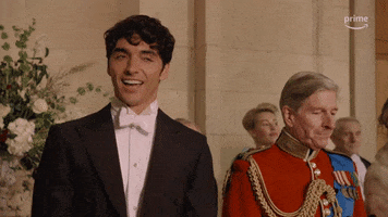 TV gif. Taylor Zakhar Perez as Kaz in Red, White, and Royal Blue. He's wearing a tux at a formal event giving a wide fake smile like he's delighted or acting surprised. 