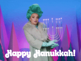 Video gif. Woman wearing a turquoise-colored wig in an elegant updo, tan fur coat, and teal gloves delicately holds up a Hanukkah menorah, striking a seductive pose. Text, "Happy Hanukkah."