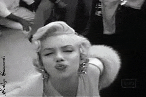 Video gif. Black and white overhead video of Marilyn Monroe wearing a white blouse, gold earrings, and a white fur shawl as she looks up at us, scrunches her nose, and blows kisses over and over.