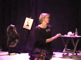 Video gif. Woman sitting on a stage with dirty feet laughs to the side, then looks at us and says, "who, me?"