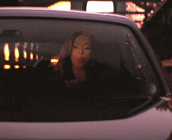 Celebrity gif. Bardi B drives toward us and sings, nodding her head rhythmically as police car lights chase behind her.