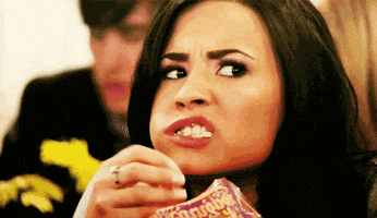 Celebrity gif. Demi Lovato is hunched over a bag of popcorn. She shovels handfuls of popcorn into her mouth and munches messily as popcorn falls out of her mouth. She glares through the corner of her eye as if captivated. 