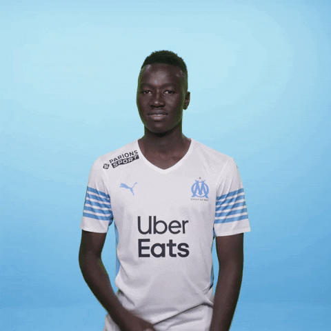 Sports gif. Pape Gueye looks down at his jersey and points to the Olympique de Marseille, smiling.