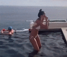 Video gif. Group of people are enjoying time in a gorgeous infinity pool that outlooks the ocean and the camera pans to two dudes in the corner. One dude is making a suction noise with his finger and his bellybutton and both are cracking up.