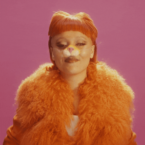 Video gif. Woman wearing an orange lion nose, furry orange jacket, and an orange wig with bangs leans in and smiles at us, giving two thumbs up against a dark pink background. Text, "Double thumbs up."