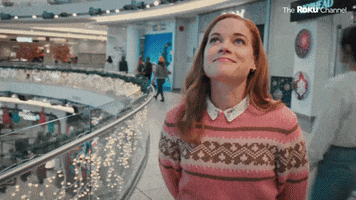 TV gif. Jane Levy as Zoey Clarke in Zoey’s Extraordinary Christmas stands in a mall with her hands behind her back. She looks up and shakes her head with a big satisfied smile on her face.