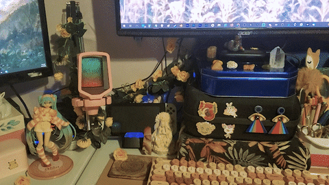 a very decorated and cozy desk setup, full of earthy and pastel colors.  you can see two monitors powered on with flowery fantasy wallpapers.  a full pen holder on the right next to a stream deck, crystals and various animal figurines decorating all surfaces of the deck. there's a nintendo switch and anime figurine of miku in the background. an aesthetic, old-fashioned looking keyboard is in the foreground, but the center piece is the plugged in, assembled and lit-up fifine microphone, now adorned with fake peachy-colored flowers as decoration.  this animated image shows heyshadylady's arm reaching out to grab the microphone and extend the boom arm to its full reach across the desk.