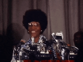 Political gif. Shirley Chisholm smiles proudly and waves at the crowd at her 1972 presidential campaign announcement as cameras flash around her. 