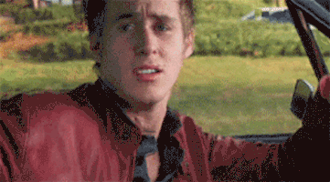 Movie gif. Driving down the street, Ryan Gosling as Richard in Murder by Numbers raises an eyebrow at us as he dances flirtatiously, then breaks into a smile.