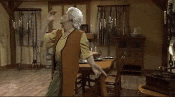 SNL gif. In an old-timey dining room with several lit candles, Willem Dafoe dances around wearing a white wig and moustache and a brown vest.