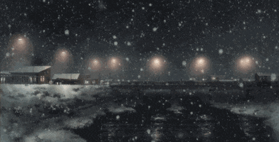Photo gif. Snow flurries fall from the night sky over a river while lights glow along a bridge through a town in the background.