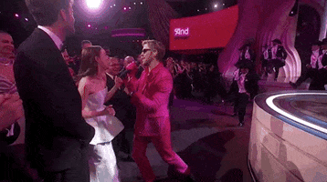 Oscars 2024 GIF. Ryan Gosling, dressed in a Barbie pink tuxedo and sunglasses, puts a microphone in the face of Emma Stone, who grabs it and fervently sings, “I’m just Ken!”