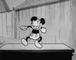 Disney gif. A retro black-and-white Mickey Mouse with a toothy smile, kicking and waving his arms in a little dance.