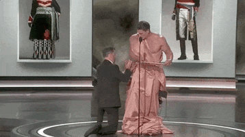 Oscars 2024 GIF. Jimmy Kimmel is kneeling down in front of John Cena as he helps Cena adjust the robe made out of a curtain that Cena has put on. Finally clothed, Cena opens the envelope and says, "And the Oscar goes to..."