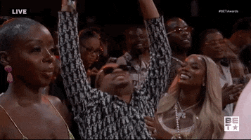 TV gif. A member of the crowd at the BET Awards throws his head back, shaking it around, and raises his arms up high like he’s praising jesus. One girl holds onto him while smiling. Another girl that’s next to him stands very still, not amused by him. 