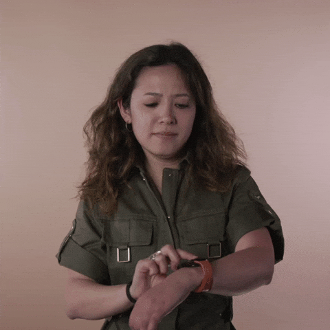 Reaction gif. A Disabled Vietnamese-American woman hemorrhagic stroke survivor with left-sided hemiplegia, brown sun-bleached hair styled in waves taps her wrist watch, looking around, brow furrowed.